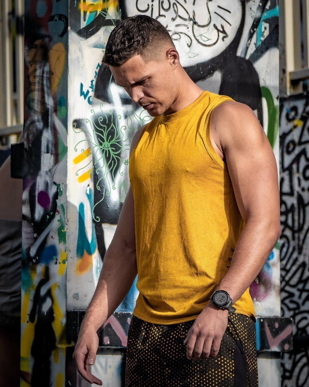 Ludovic, coach sportif, en débardeur jaune devant un mur couvert de graffitis.